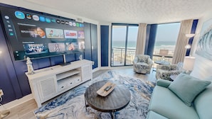 Una habitación de hotel moderna con un televisor grande que muestra varios servicios de streaming, un balcón con vista al océano y una zona de estar acogedora con un tapete con estampado de mármol.