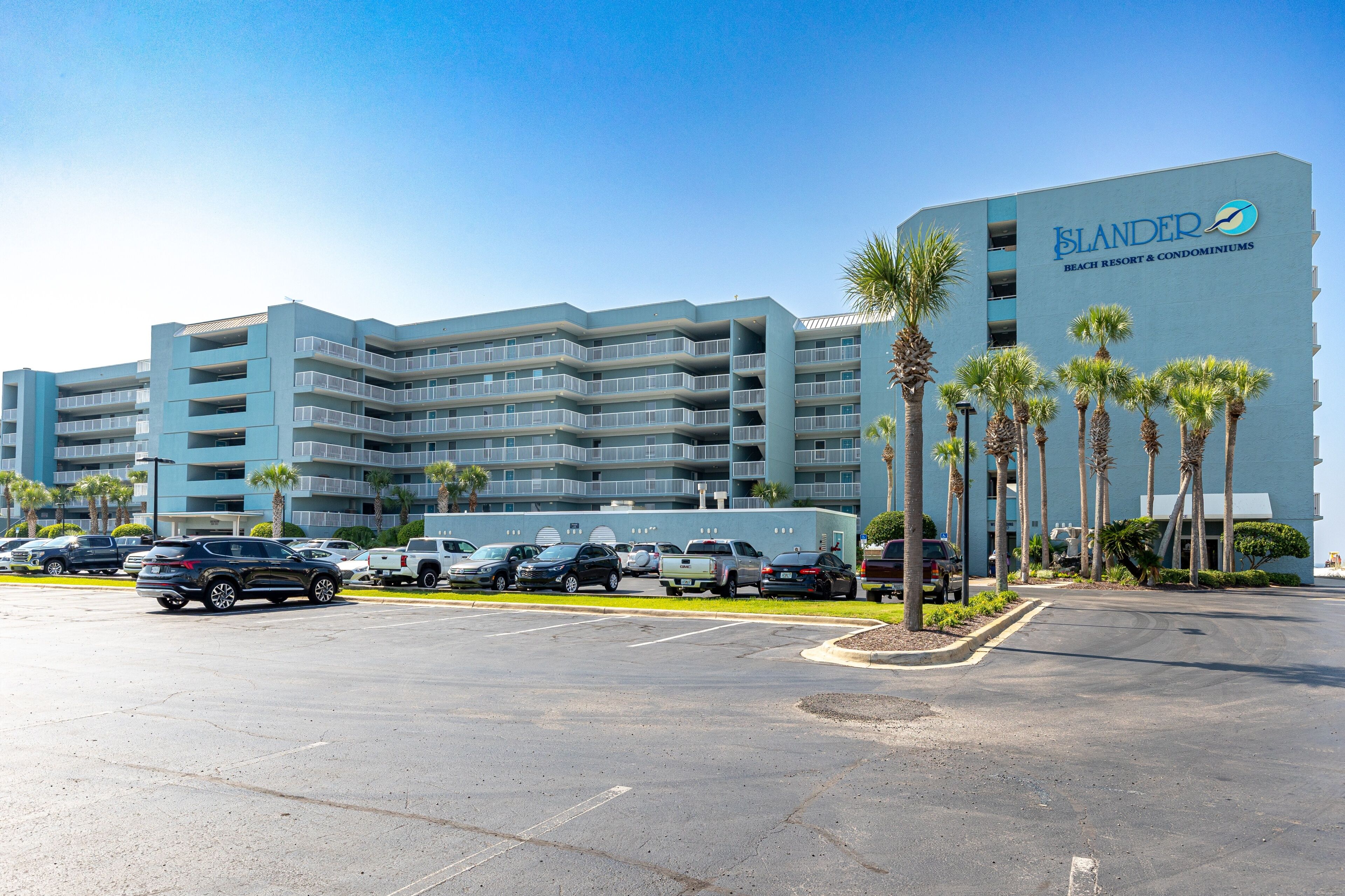 A multi-story hotel with a blue facade, named 'SLANDER BEACH RESORT & CONDOMINIUMS'. It features a parking lot with several cars and palm trees in front.