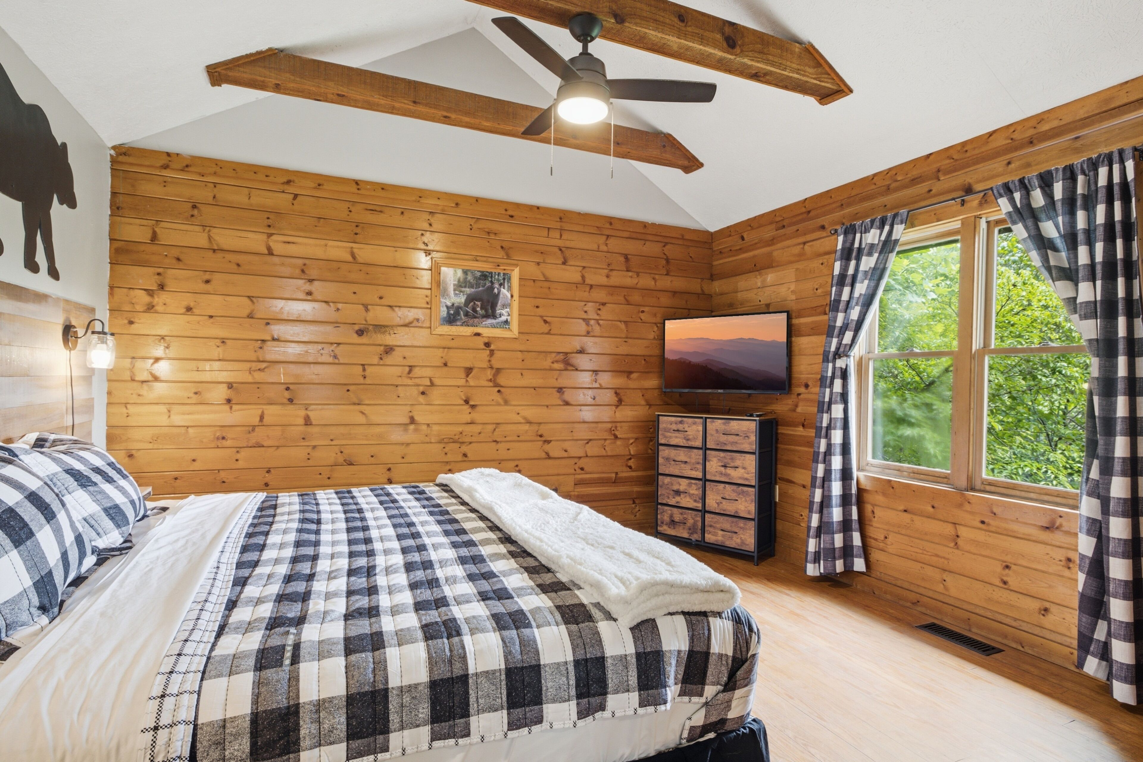 Un dormitorio con paneles de madera, una cama con colcha a cuadros, un ventilador de techo, un televisor y una ventana con cortinas.
