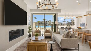 A modern living room with a flat-screen TV, a chandelier, a sofa, and a dining area with a view of a pool. - Barefoot Beach House A 3 Bedroom Townhouse (St. George)