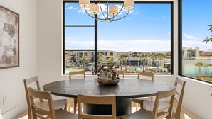 A dining area with a round table, chairs, a chandelier, and a large window with a view of a pool and buildings. - Barefoot Beach House C 3 Bedroom Condo (St. George)