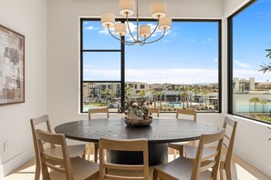 A dining area with a round table, chairs, a chandelier, and a large window with a view of a pool and buildings. - Barefoot Beach House C 3 Bedroom Condo (St. George)