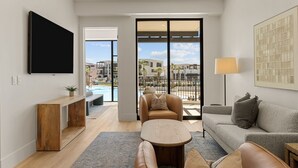 Una sala de estar moderna con un televisor de pantalla plana, una consola de madera, un sofá, una mesa de centro y vistas a una piscina y edificios.