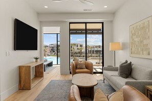 Ein modernes Wohnzimmer mit einem Flachbildfernseher, einem Holzkonsolentisch, einer Couch, einem Couchtisch und Blick auf einen Pool und Gebäude.