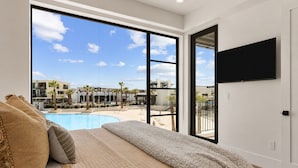 Un dormitorio con un ventanal grande, una cama, un televisor y vistas a una piscina y edificios.