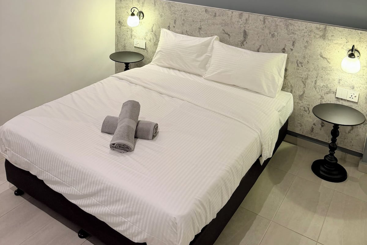 A neatly made bed with white linens and rolled towels, two bedside tables with lamps, a grey textured headboard, and a white air conditioning unit on the wall.