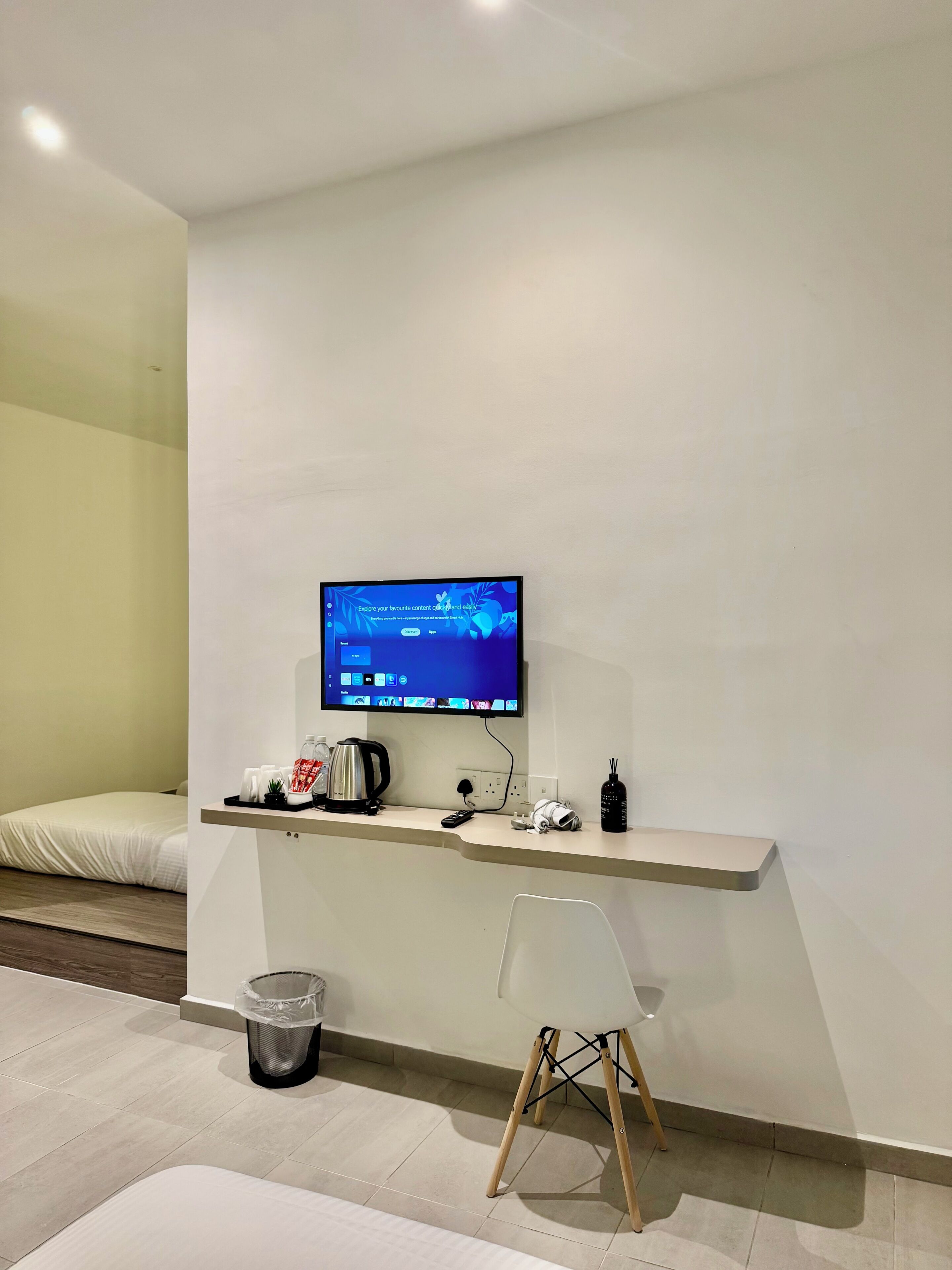 A modern hotel room with a flat-screen TV mounted on the wall, a small desk with a kettle, a trash bin, and a chair.
