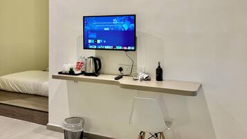 A modern hotel room with a flat-screen TV mounted on the wall, a small desk with a kettle, a trash bin, and a chair.