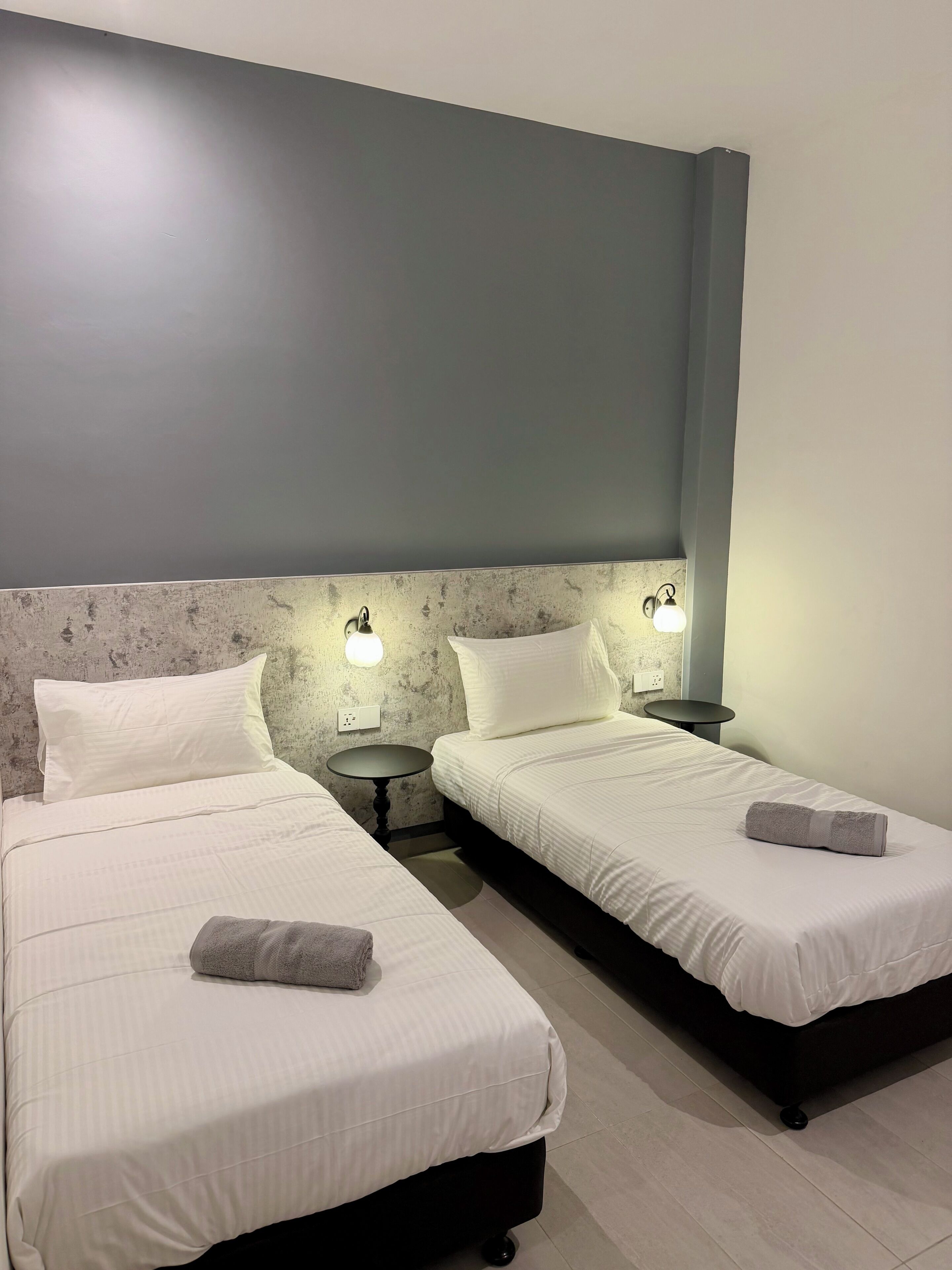 Habitación de hotel con dos camas, cada una con una almohada y una toalla, una mesita redonda pequeña y luces fijadas a la pared.