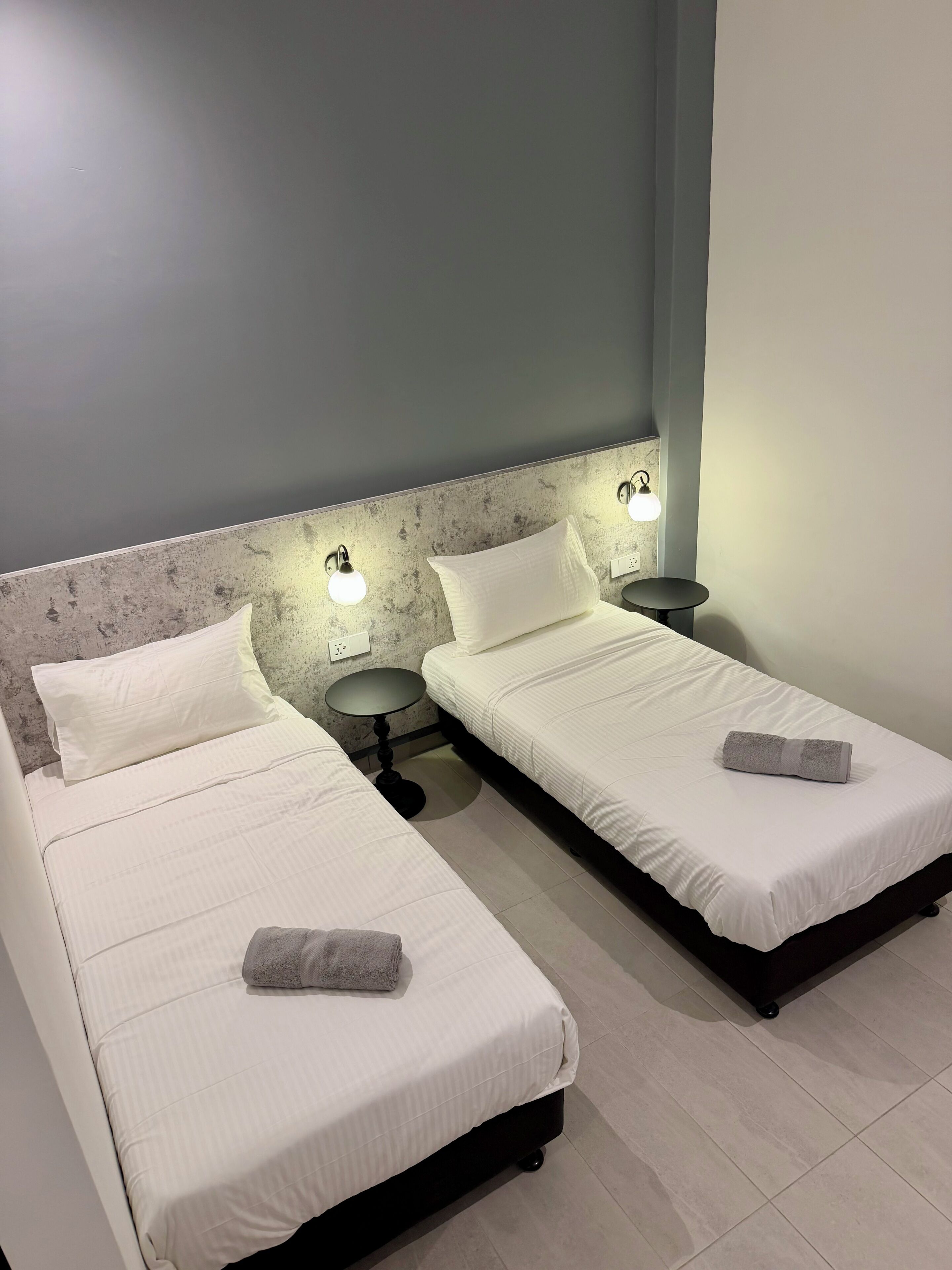 Dos llits individuals amb roba de llit blanca, una taula rodona petita i llums fixades a la paret en una habitació amb un mur sense decoració i terra d'alzapa.