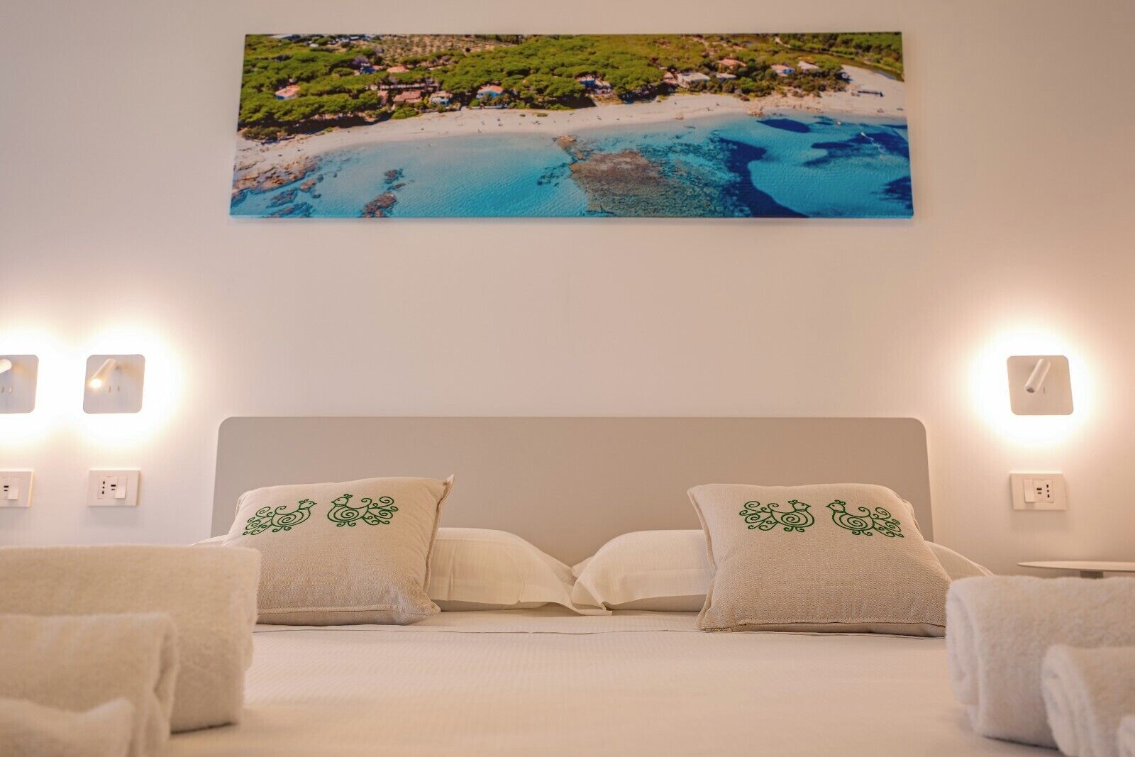 A bedroom with a bed, two pillows with green designs, a wall-mounted picture of a beach, and bedside lamps.