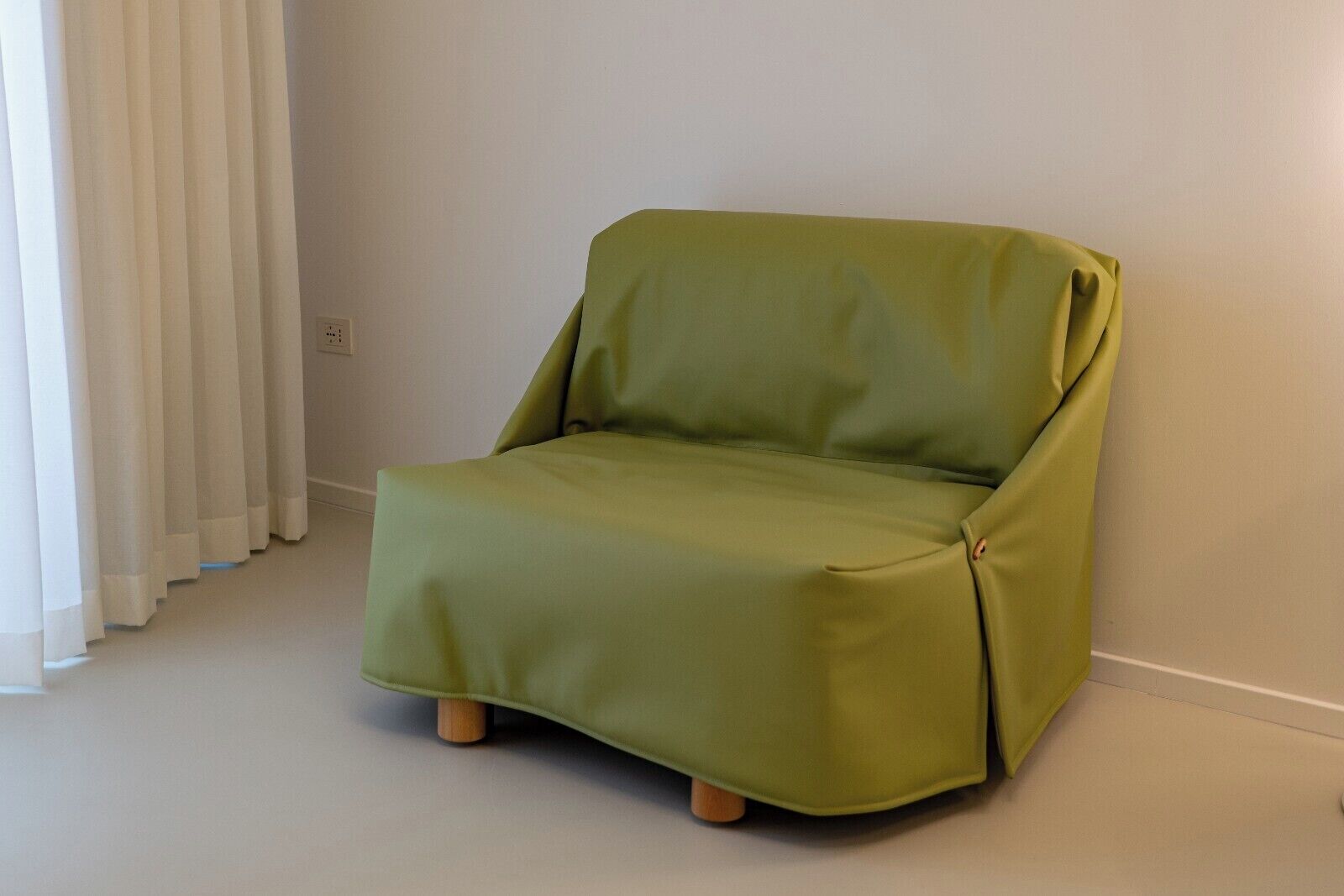 Un fauteuil vert aux pieds en bois, dans une pièce aux murs et au sol blancs.