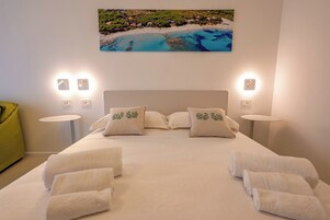 Habitación de hotel con una cama, dos mesitas de noche, una silla verde y un cuadro de una playa.