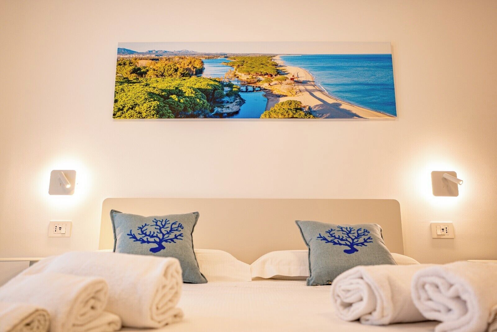 Un dormitorio con una cama, dos almohadas con diseños de árboles, un cuadro de un paisaje costero y dos lámparas de pared.