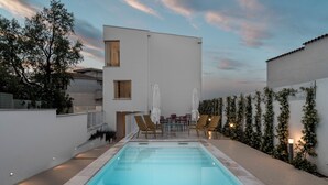 Una piscina con sillas de descanso y una mesa, rodeada por un edificio blanco y follaje.