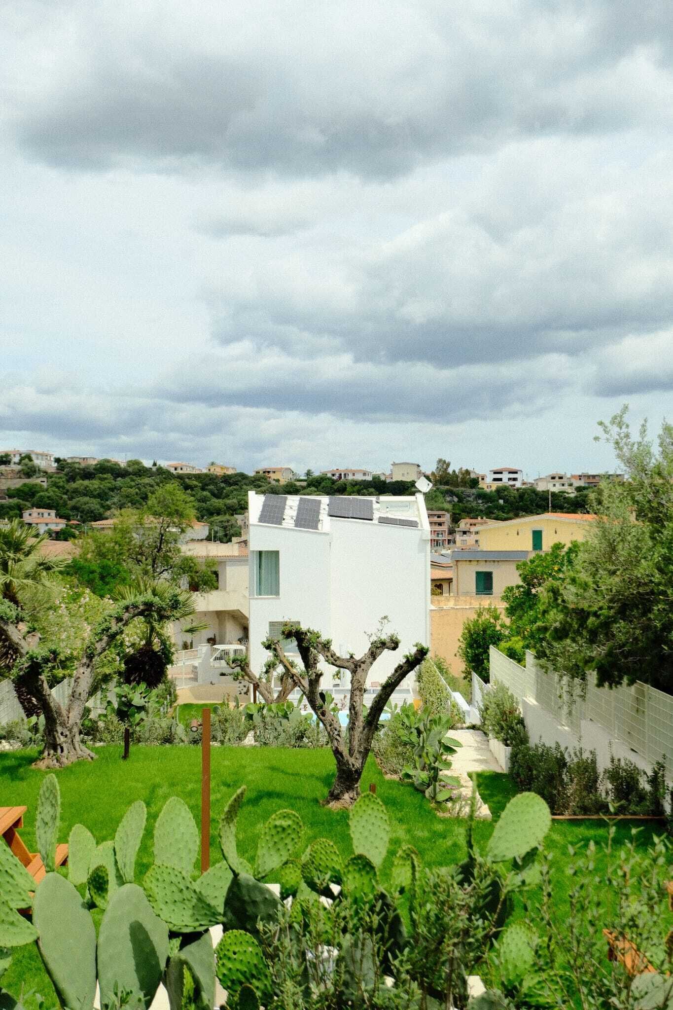 Un barrio residencial con un edificio moderno blanco, vegetación verde y un cielo nublado.