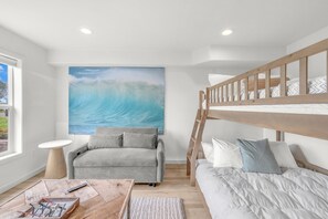 Ein Zimmer mit einem Etagenbett, einer Schlafcouch, einem Wellenbild, einem kleinen Tisch und einem Fenster.