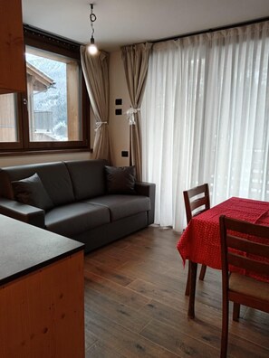 A room with a sofa, a dining table with chairs, a window with curtains, and a hanging light fixture.