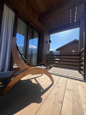 Eine Holzdielen-Terrasse mit einem Holzstuhl, großen Fenstern mit Vorhängen und Blick auf einen Berg.