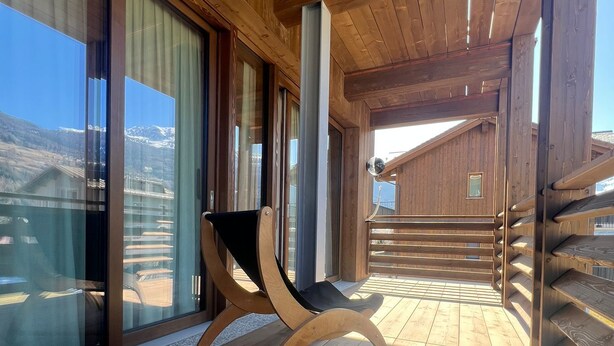 Una terraza de madera con una silla, ventanas grandes y vistas a montañas.