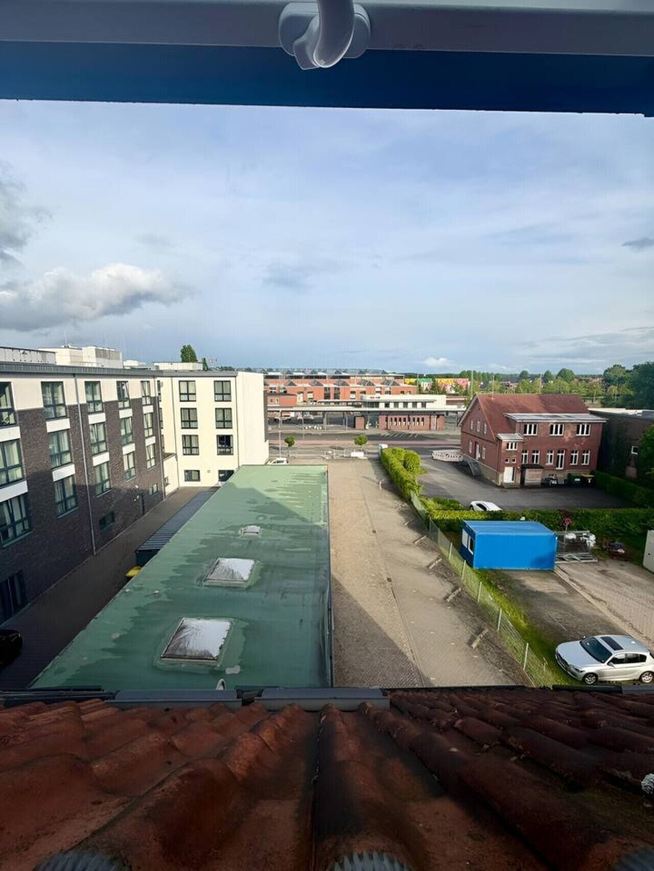 A view from a window overlooking a parking lot, a green-roofed building, and a red-roofed building with a blue container.