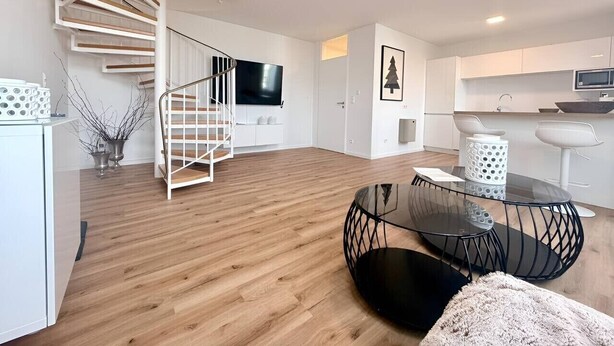 A modern living room with a spiral staircase, a flat-screen TV, a dining area with a glass table, and a kitchen with white cabinets.
