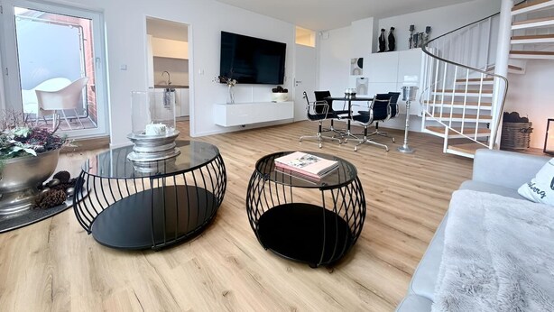 A modern living room with a glass coffee table, a sofa, a TV mounted on the wall, a dining area with chairs, and a spiral staircase. - City-Loft Lingen Apartment 3 in the heart of Lingen (Lingen (Ems))