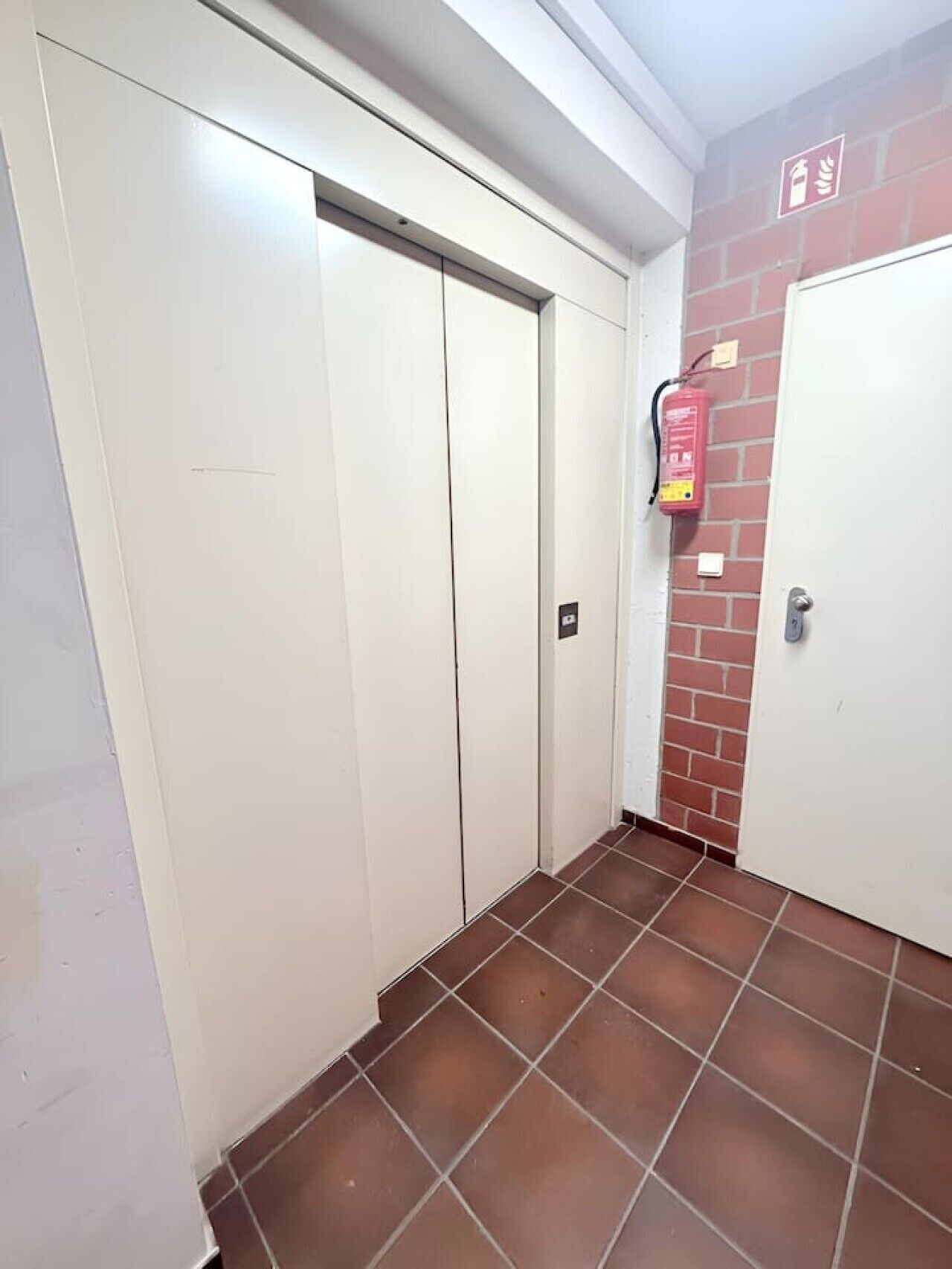 Ein Flur mit an der Wand befestigtem Feuerlöscher, einer Tür und einem gefliesten Boden.