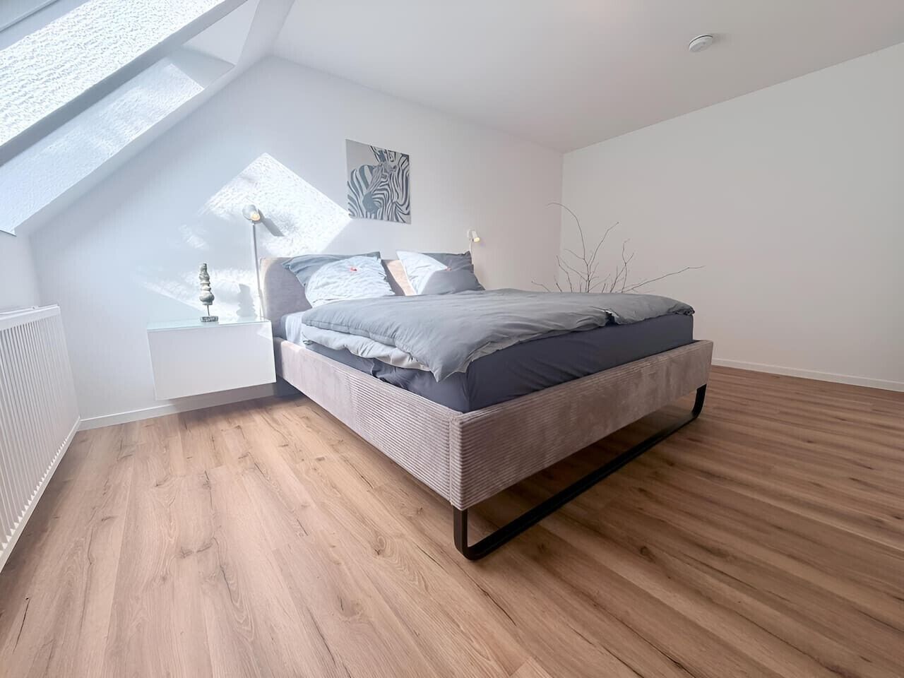 Ein modernes Schlafzimmer mit schrägverdachter Decke, einem Bett, einem Nachttisch, einer Wandleuchte und einer Heizung.