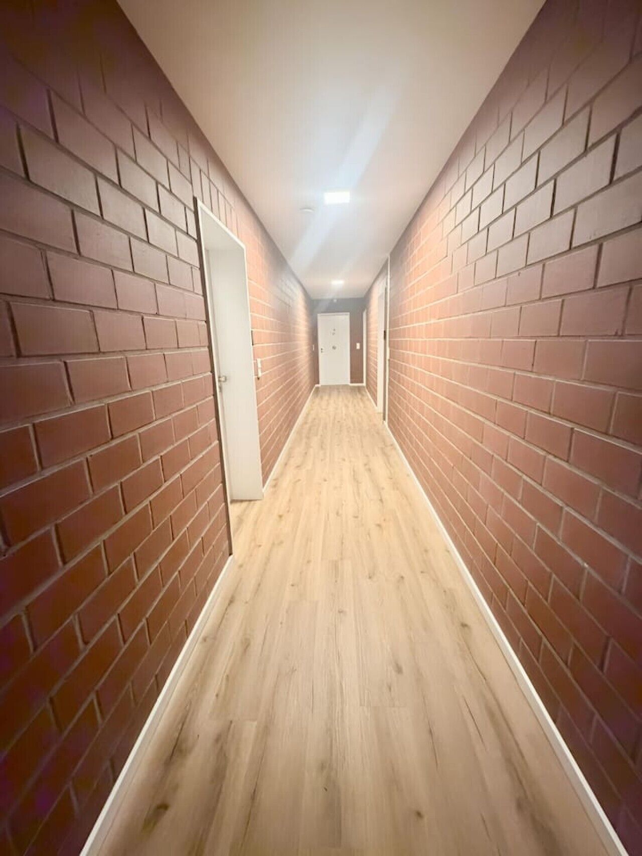 A narrow hallway with brick walls and wooden flooring, leading to a door at the end.