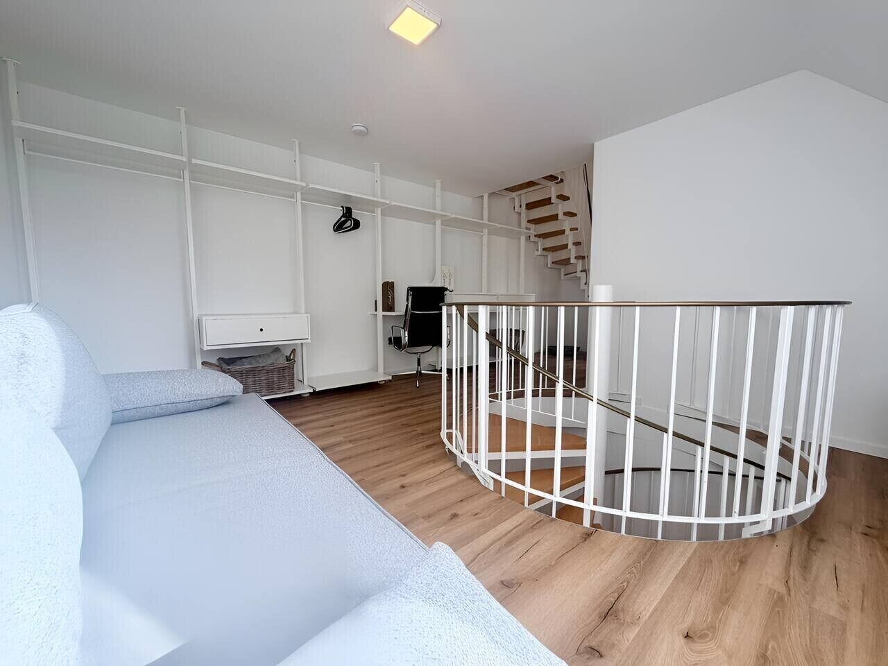 A modern interior with a white sofa, a wooden floor, a white railing, and a staircase.