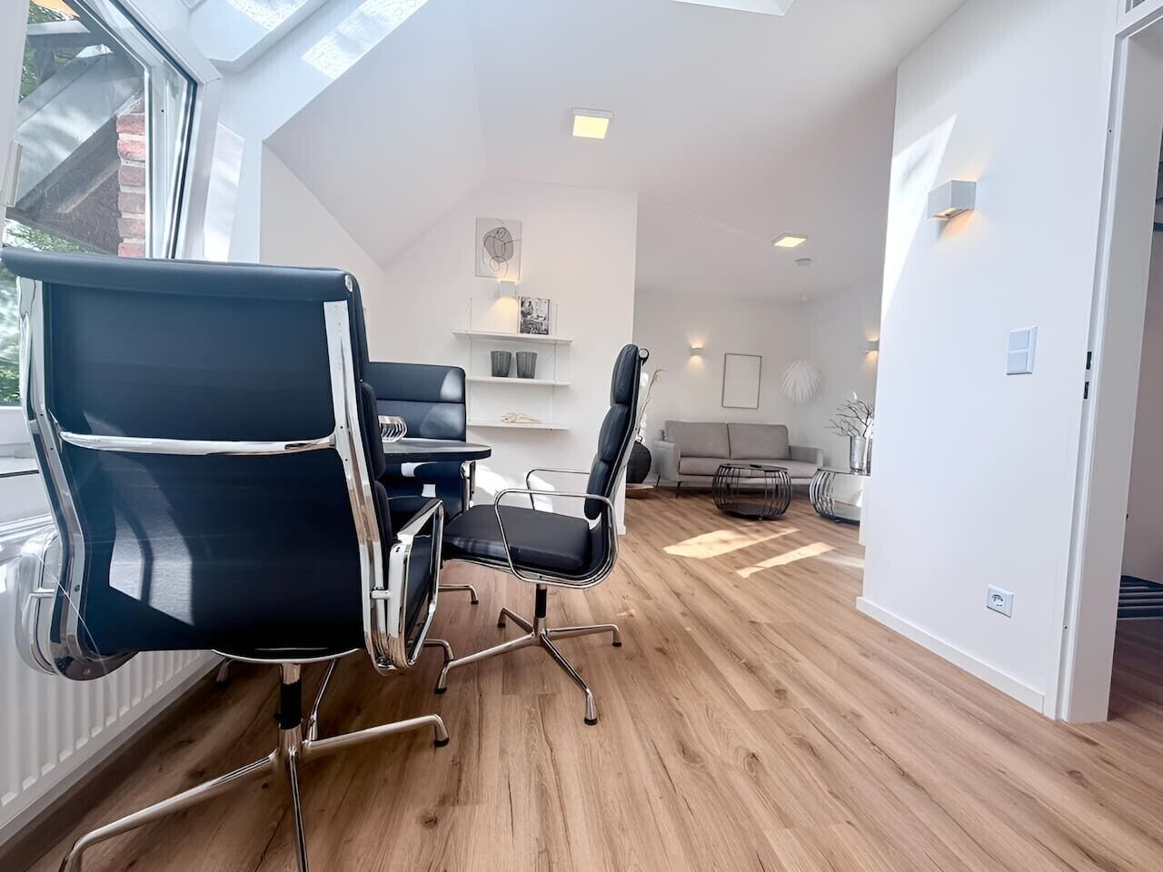 Et moderne kontor med et konferencemødebord, to sorte kontorstole, en sofa og en hvid væg med en hylde.