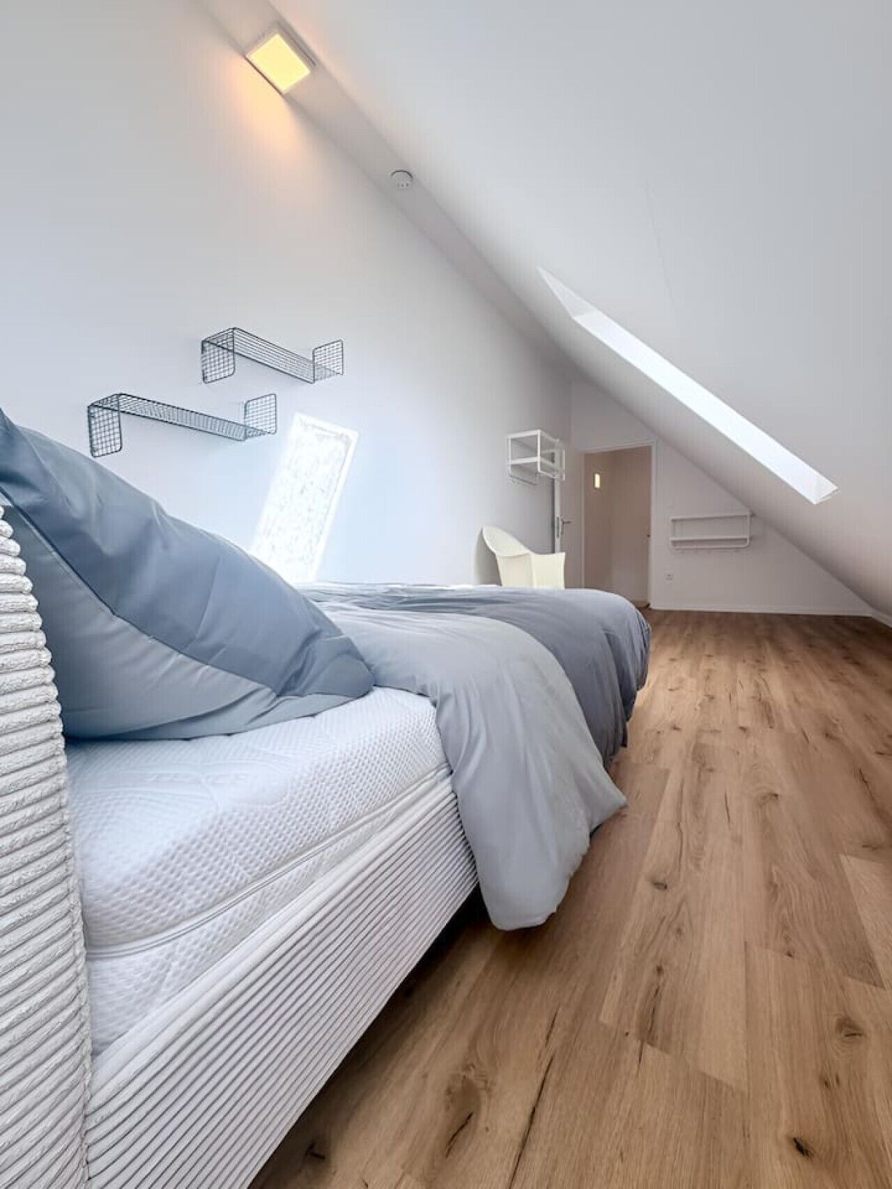 Un dormitorio tipo loft moderno con techo inclinado, cama con ropa de cama gris, una silla blanca y suelo de madera.