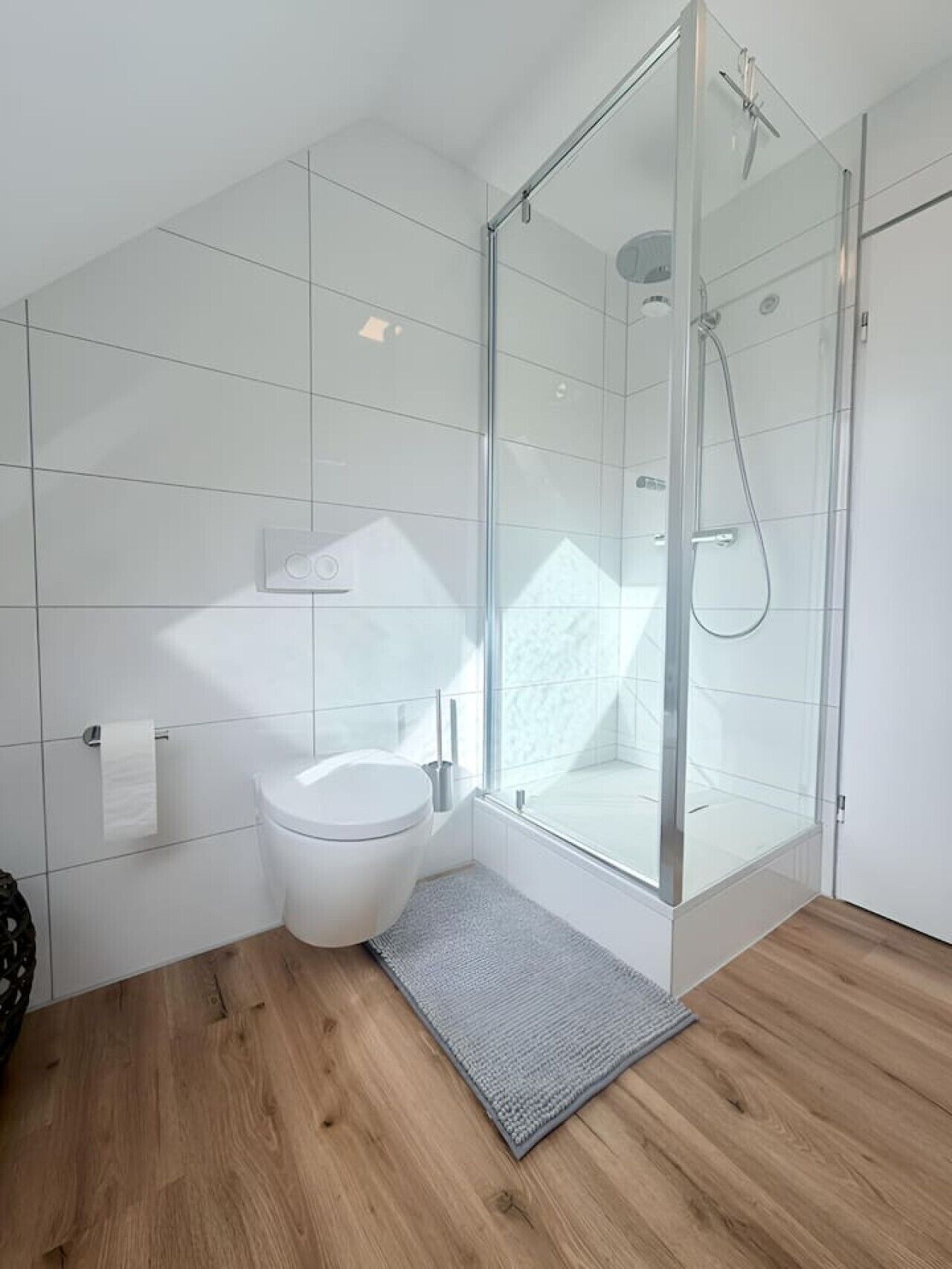 A modern bathroom with a glass shower enclosure, a white toilet, and a grey rug on the wooden floor.