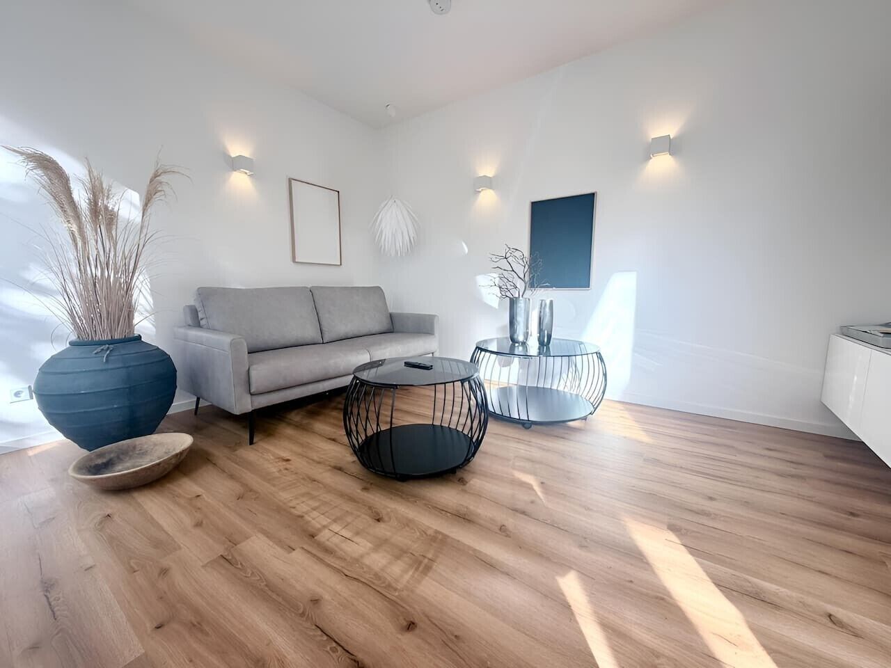 Ein modernes Wohnzimmer mit einem grauen Sofa, zwei schwarzen Tischchen aus Drahtgestell, einer großen Vase mit Trockenpflanzen und einem weißen Schrank.