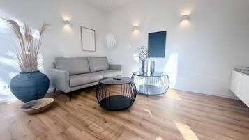 Ein modernes Wohnzimmer mit einem grauen Sofa, zwei schwarzen Tischchen aus Drahtgestell, einer großen Vase mit Trockenpflanzen und einem weißen Schrank.