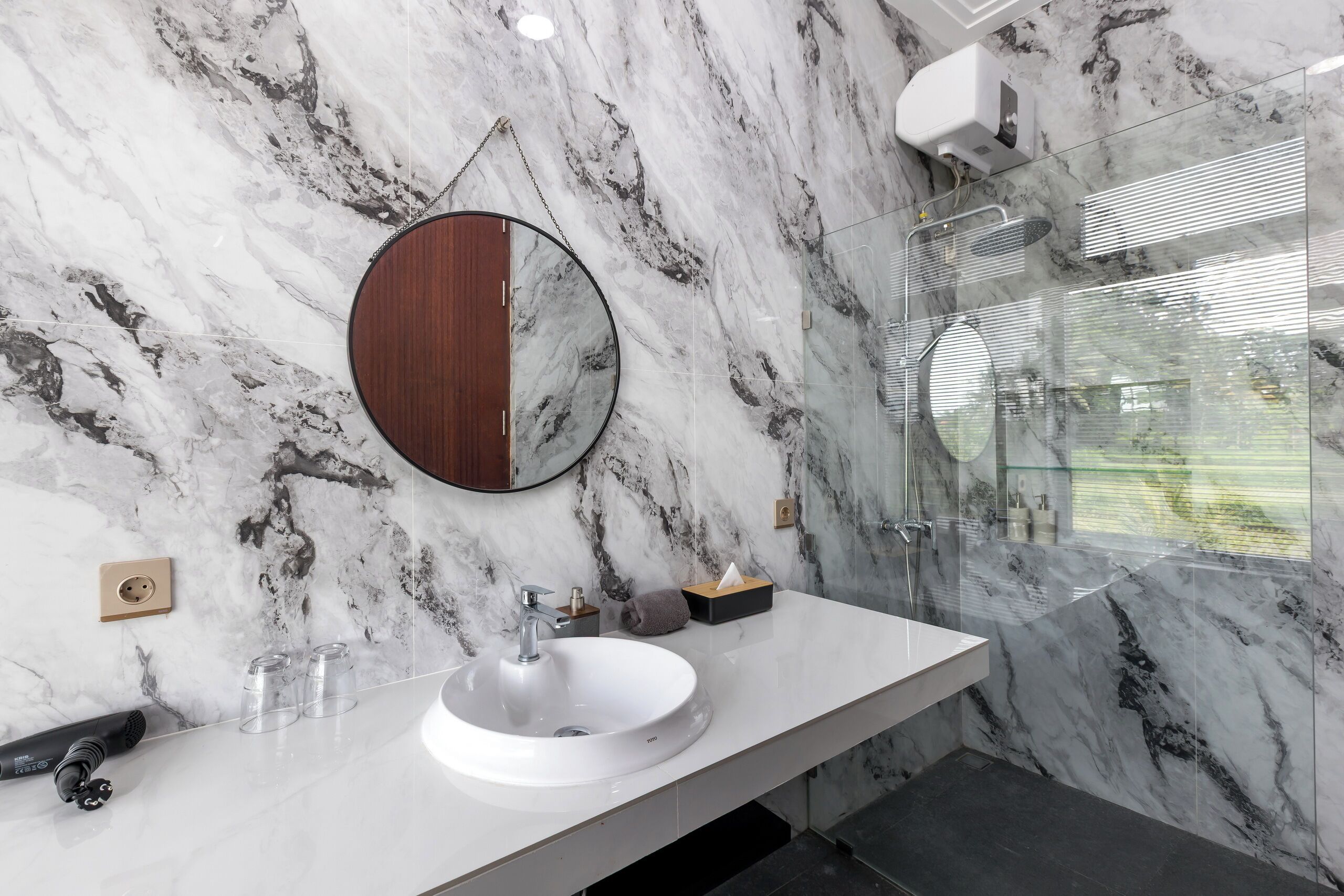 A bathroom with a marble wall, a round mirror, a white sink, and a glass-enclosed shower.