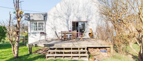 Een wit huis met een terras, een eettafel, stoelen, een houten bank en een schoorsteen.