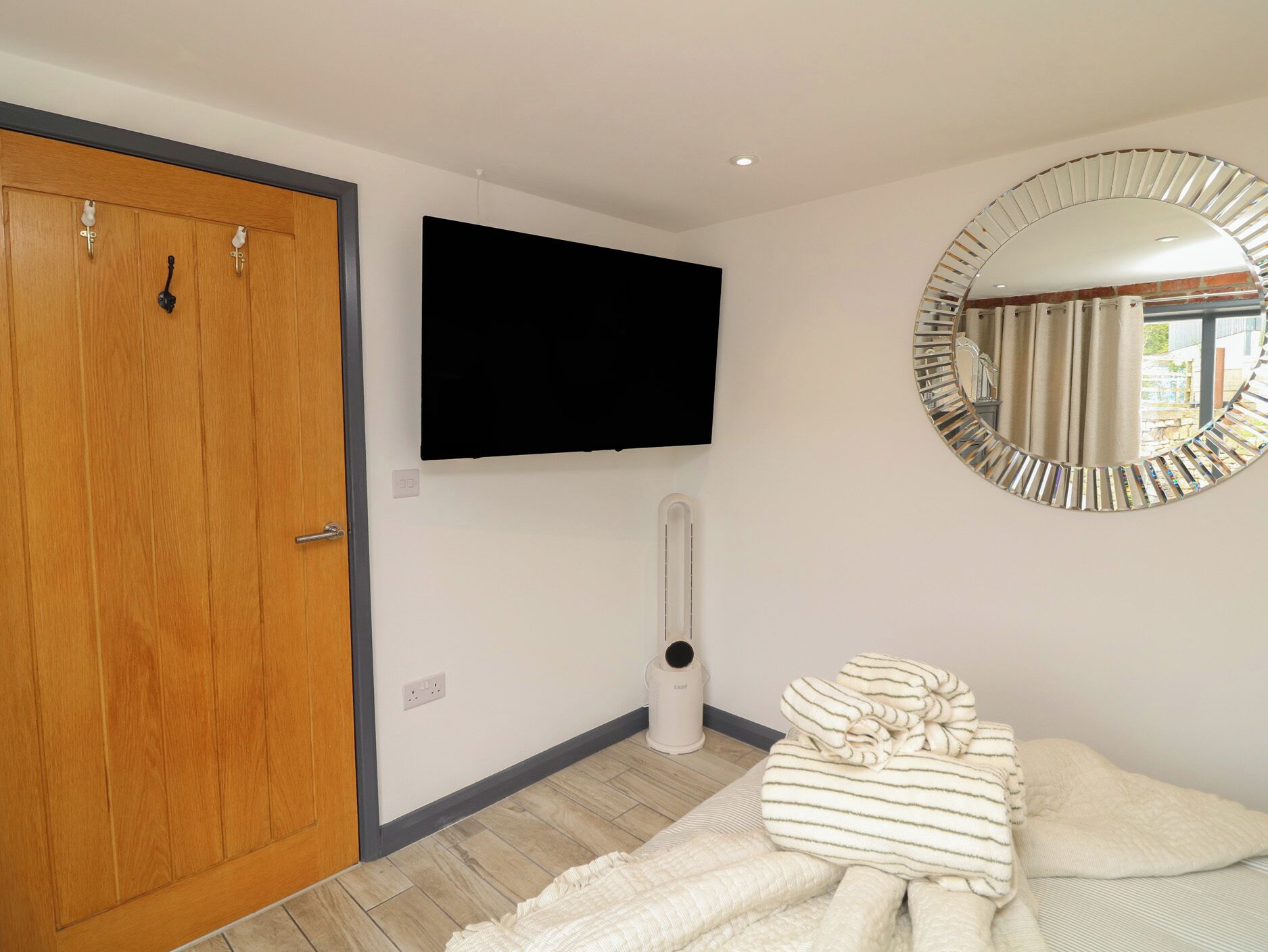 A room with a wooden door, a mounted TV, a round mirror, a radiator, and a bed with striped bedding.