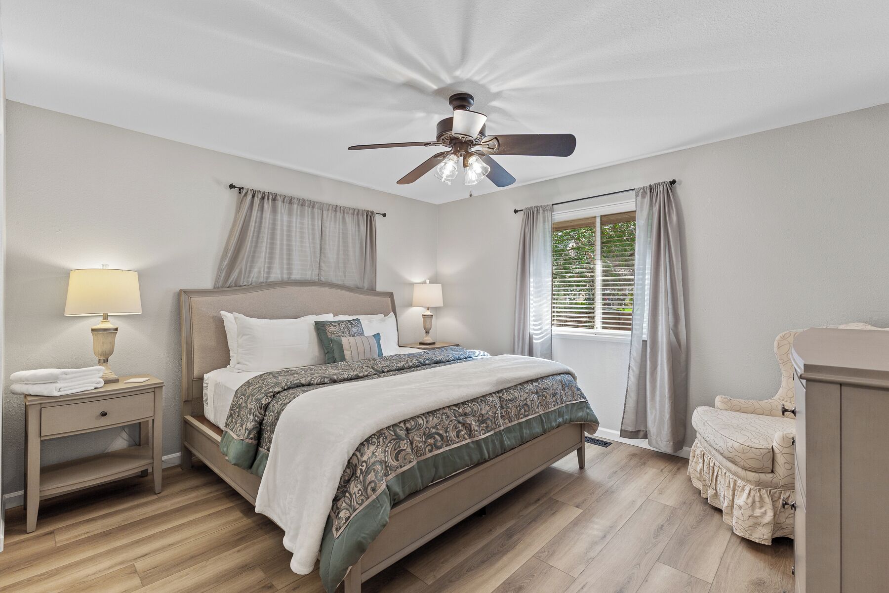 A bedroom with a bed, two bedside tables with lamps, a ceiling fan, and a window with curtains.