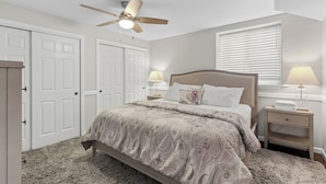 Une chambre à coucher comprenant un lit, des lampes de chevet, un ventilateur de plafond, une fenêtre avec des stores et une porte.