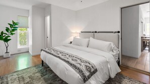 Un dormitorio con cama, ventilador de techo, ventana con persianas, mesita de noche con lámpara y una planta en maceta.