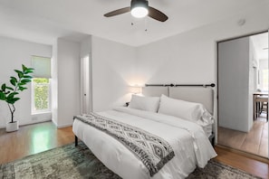 Ein Schlafzimmer mit einem Bett, einem Deckenventilator, einem Fenster mit Jalousien, einem Nachttisch mit Lampe und einer Topfpflanze.