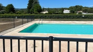 Una piscina rectangular con valla de metal negro, rodeada de vegetación y casas al fondo.