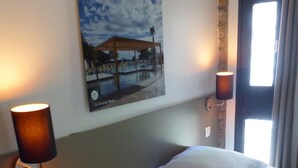 Habitación de hotel con cama, dos lámparas de pared, un cuadro de un puerto deportivo y una ventana con cortinas.