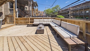 A wooden deck with a sofa and tables, a brick building, and a balcony. - The Dakin Estate: A refined 4BR Art Deco speakeasy duplex in Wrigleyville. Featuring curated interiors, elevated finishes, and a private entertainment lounge just minutes from Wrigley Field, dining, nightlife, and Chicago’s iconic attractions. (Chicago)