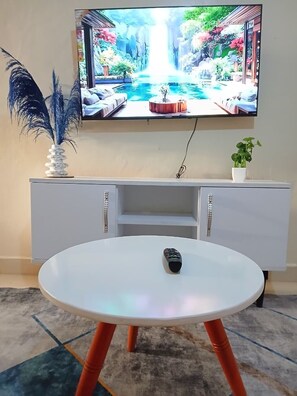 A living room with a TV mounted on the wall, a white entertainment unit, a round table with a remote control, and a potted plant. - Beautiful and cozy odern bedroom unit in Mirema, Nairobi. 20 min drive from CBD. (Nairobi)