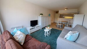 Ett vardagsrum med en brun soffa, en grå soffa, en tv på ett vitt skåp, ett litet bord med en växt och ett köksområde i bakgrunden.