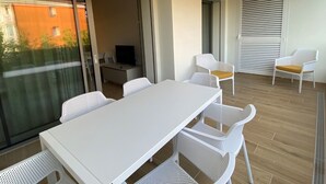 Un balcón con una mesa y sillas blancas, un televisor y una puerta corrediza.