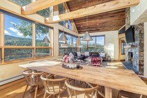 Ein geräumiges Zimmer mit einem Holztisch, Barhocker, einem Kamin und großen Fenstern mit Blick ins Freie.